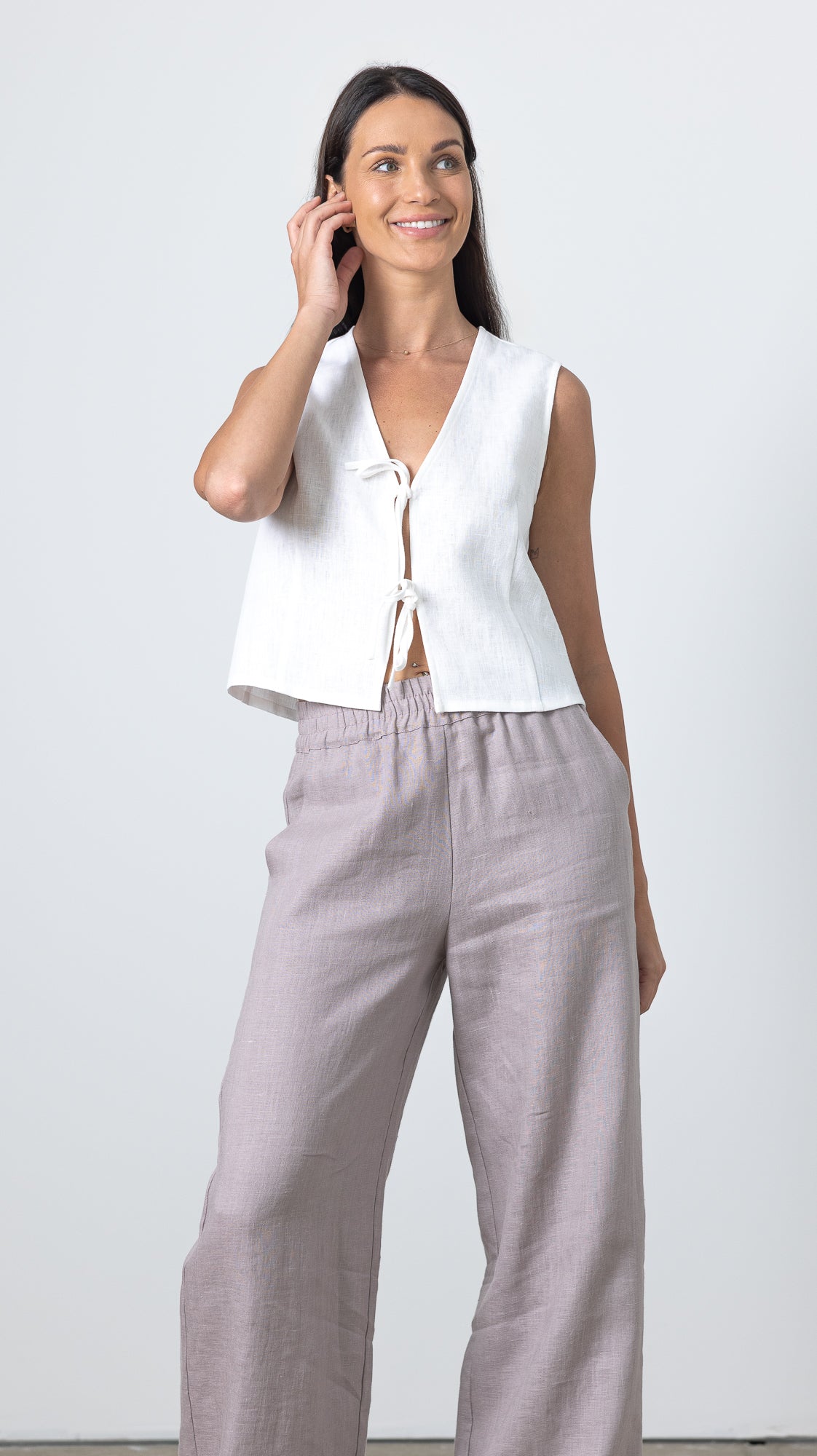 Woman touching her hair while wearing a white linen tie front top and relaxed linen trousers, minimalist linen fashion