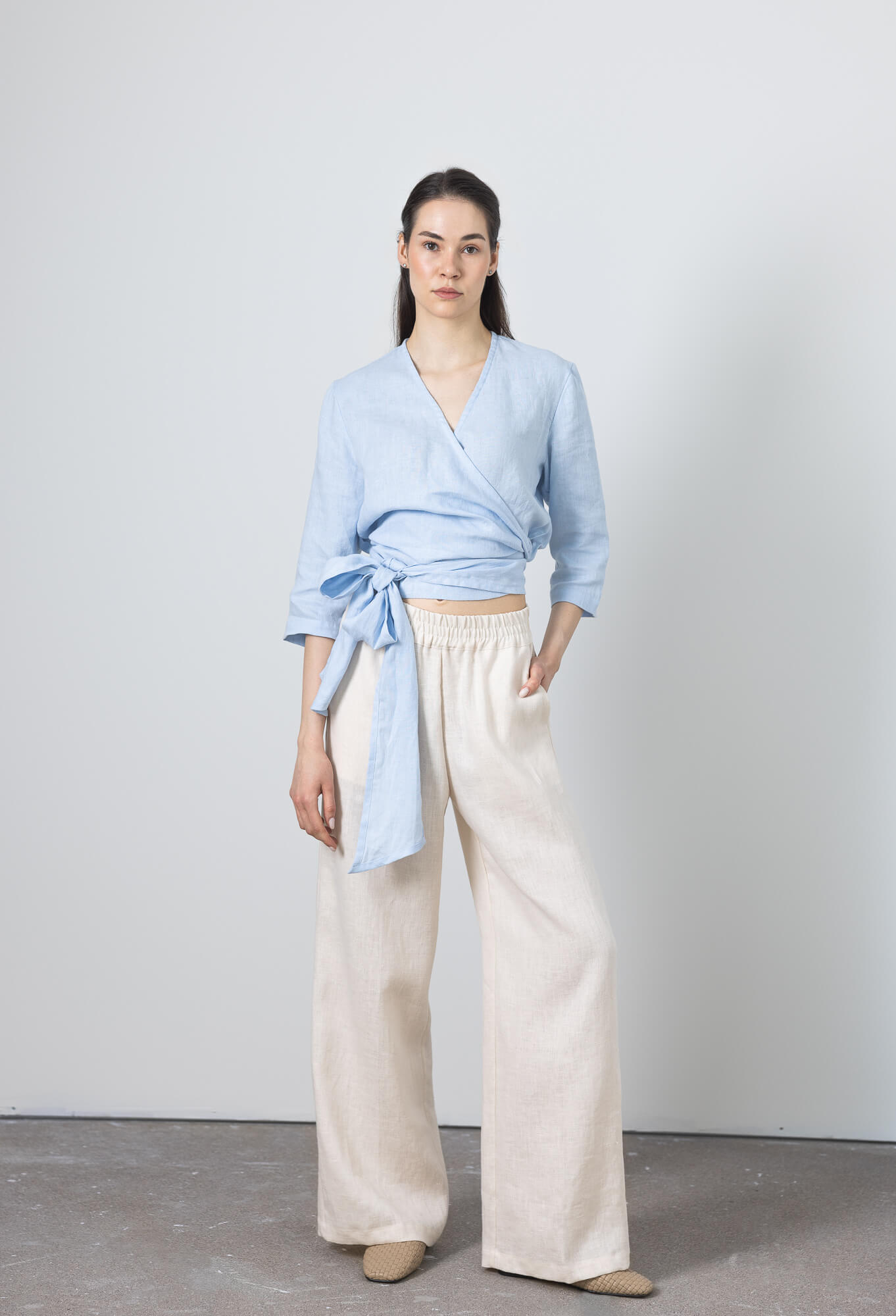 Elegant linen wrap blouse with bow detail styled with neutral wide-leg linen pants, worn by a model in a bright studio setting